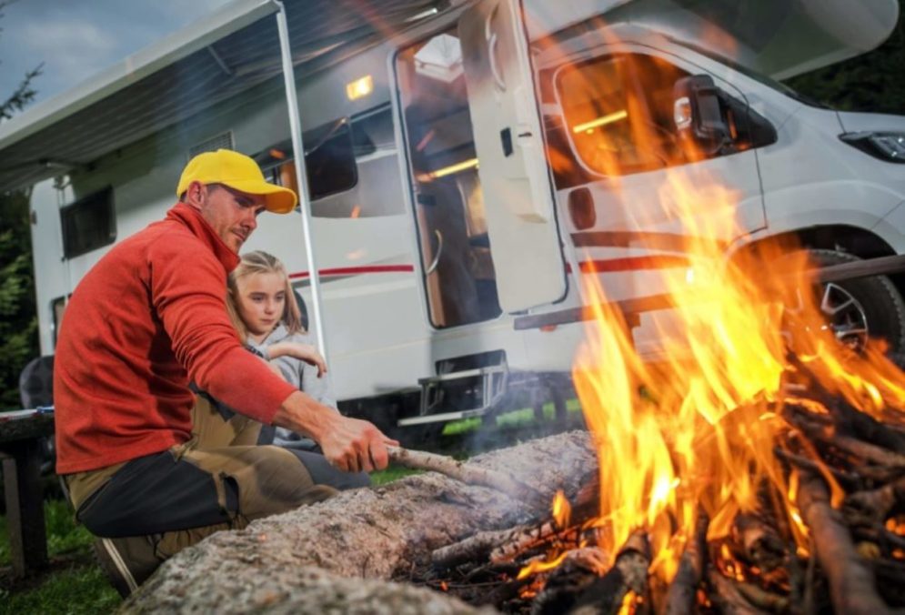 A father and daughter stoke a fire next to their RV camper.